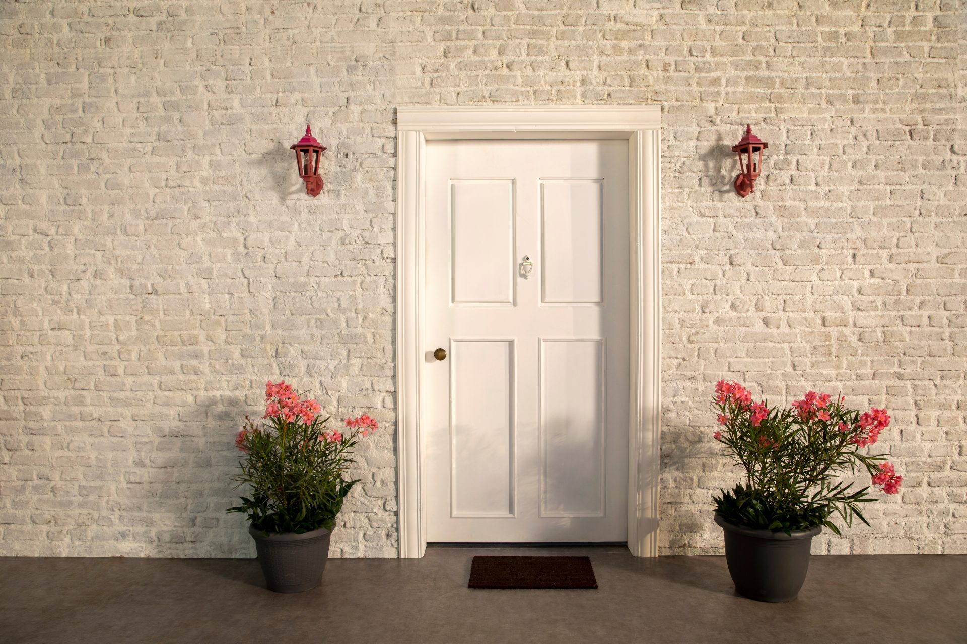A classic white paneled front door is flanked on either side by blooming planters and decorative light fixtures