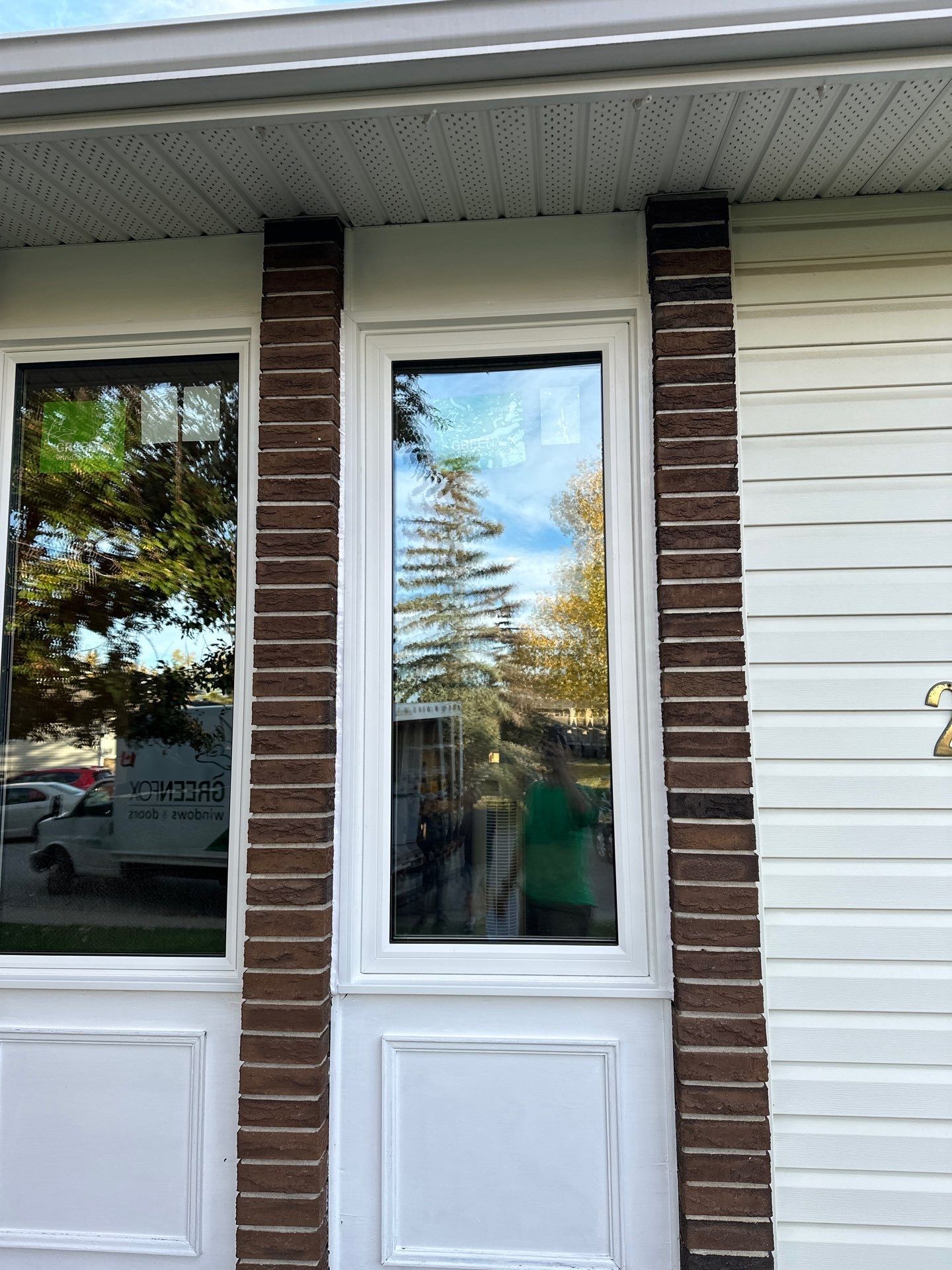 A tall, narrow replacement window with a white frame, installed between two brown brick columns and surrounded by white siding