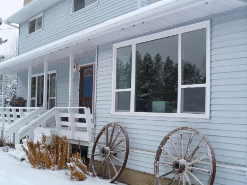 A light blue two-story house with white trim and a snow-covered porch, featuring large front windows and decorative wagon wheels