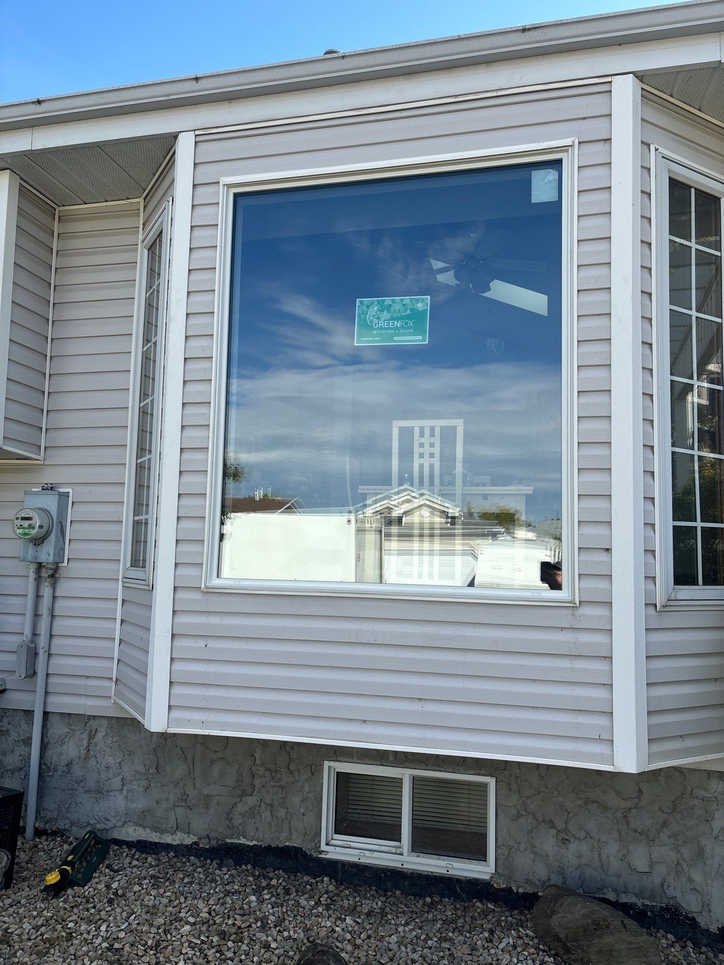 A large picture window with a GreenFox sticker installed on a beige vinyl-sided home in Camrose, AB, reflecting a blue sky and nearby rooftops.