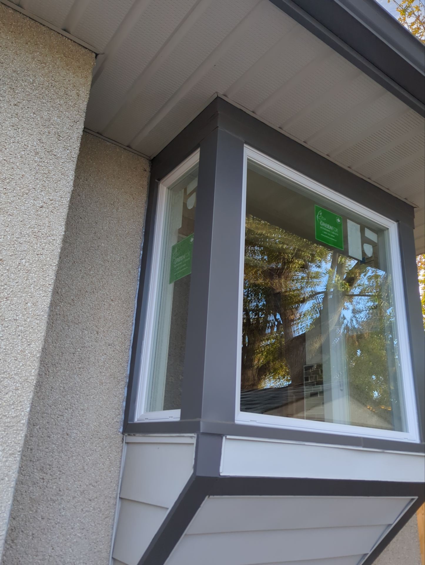 A close-up view of a newly installed bay window with a white frame and dark gray trim on a textured beige and gray exterior wall