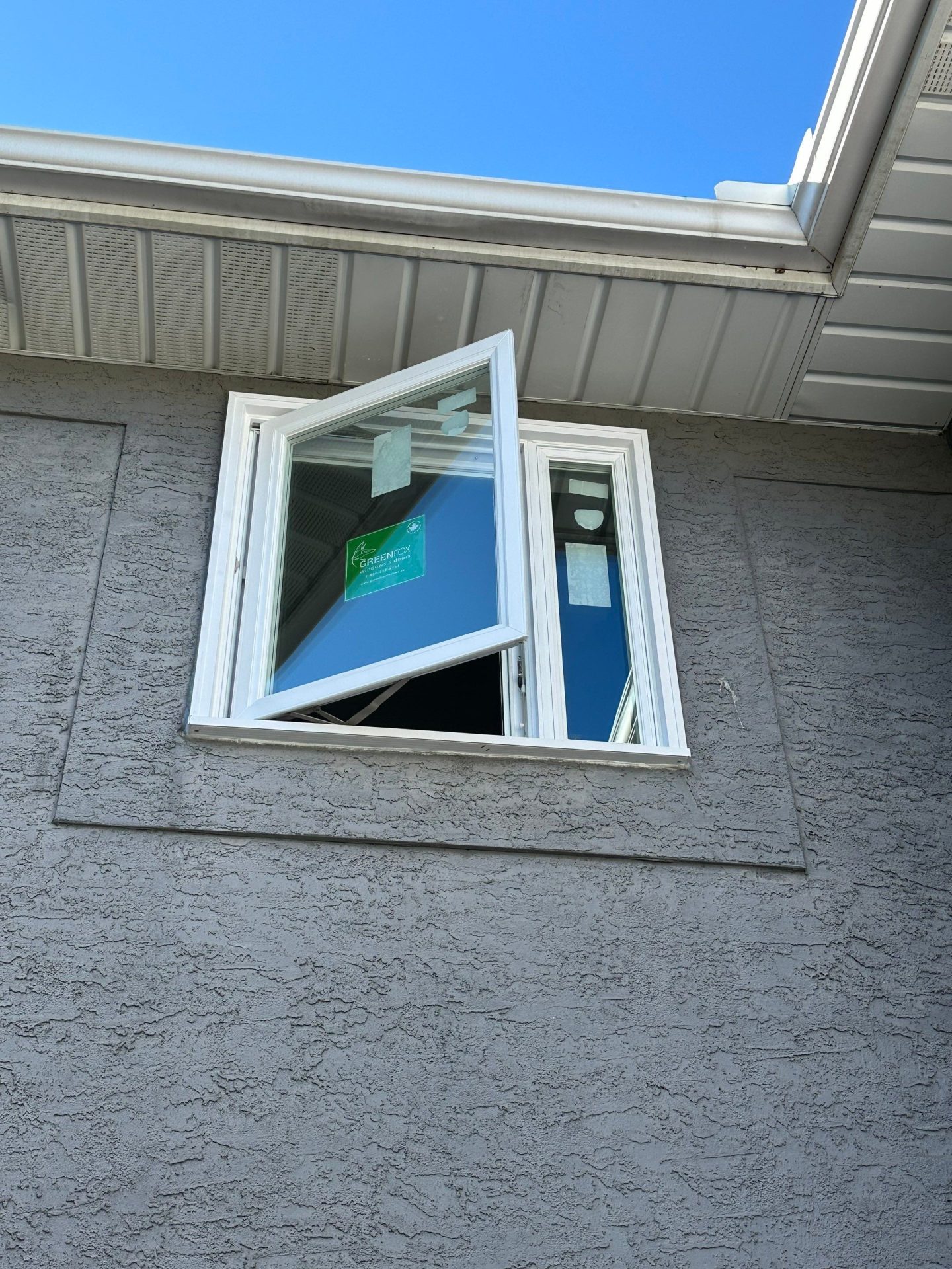 A single open casement window with a white frame is set into the upper floor of a gray stucco house, with a bright blue sky in the background