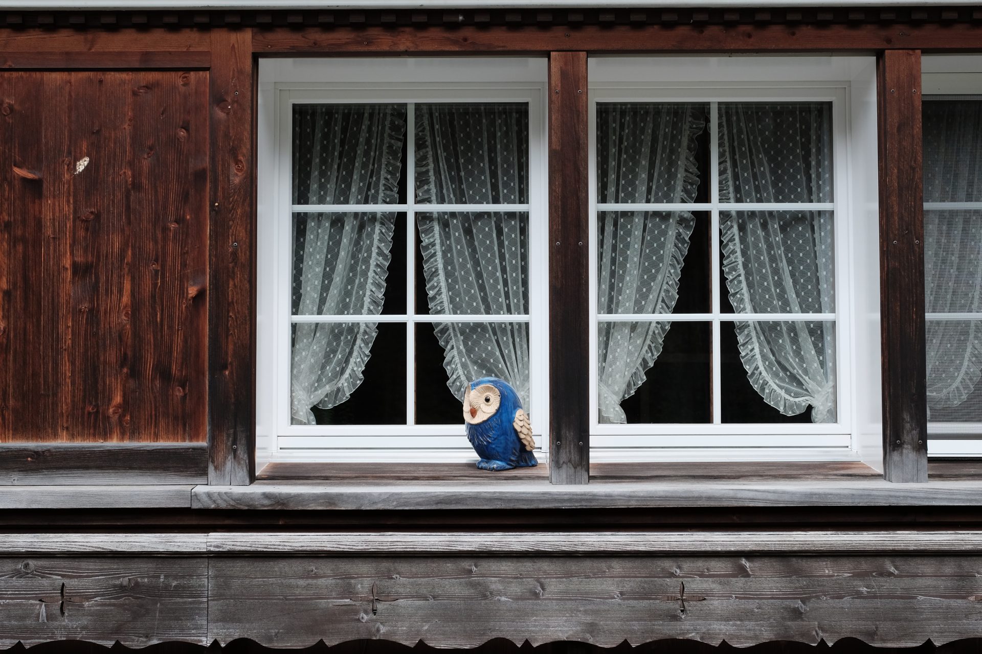 White windows on a wood house with curtains inside and a decorative owl on the window ledge.