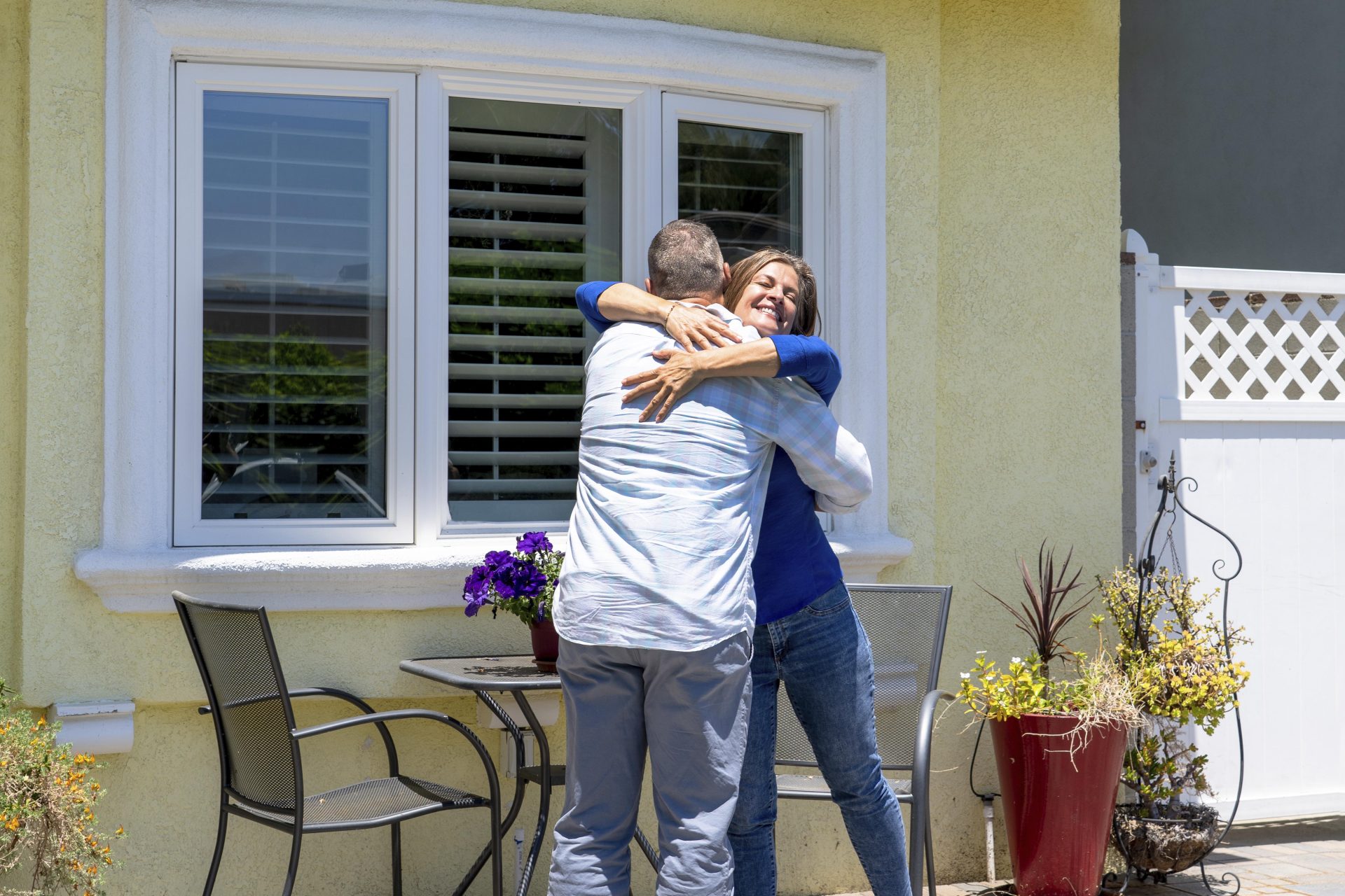 A smiling woman hugging a man in front of a stucco house with white bay windows