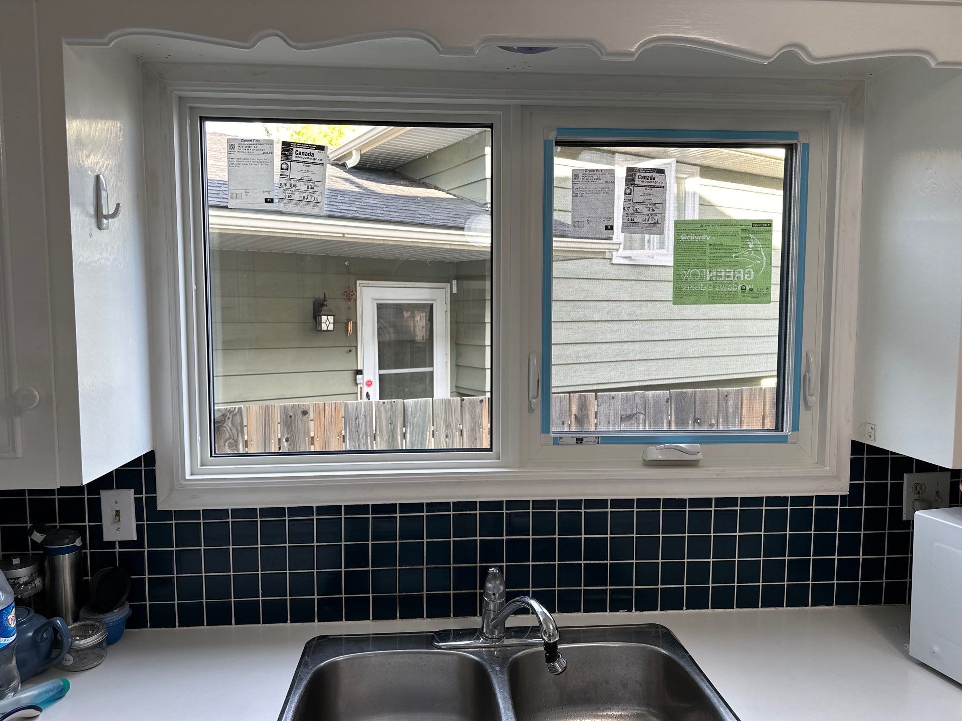 An inside view of casement windows over a kitchen sink, looking outside at a wood fence anda  neighbor’s house