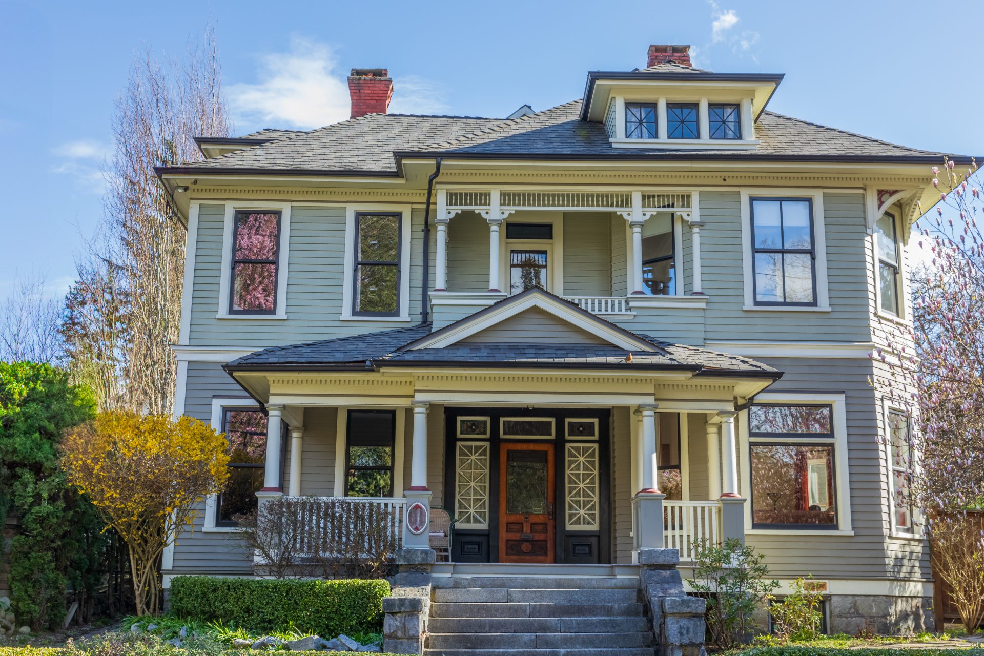 A multi-story Victorian-style tan house with dark windows, a wooden front door, and an asphalt shingle roof.