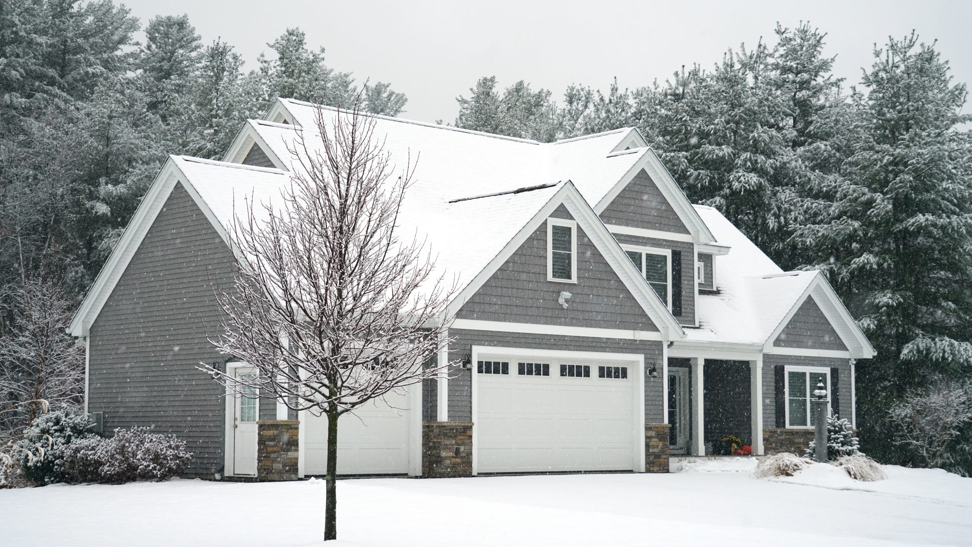 Large gray two-story home surrounded by snow-covered trees with white trim, stone accents, and a snow-covered roof during winter snowfall