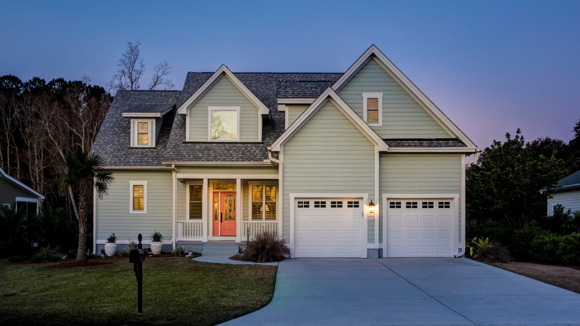 Two-story home exterior at dusk with soft green siding, white trim, and illuminated windows beside a double garage