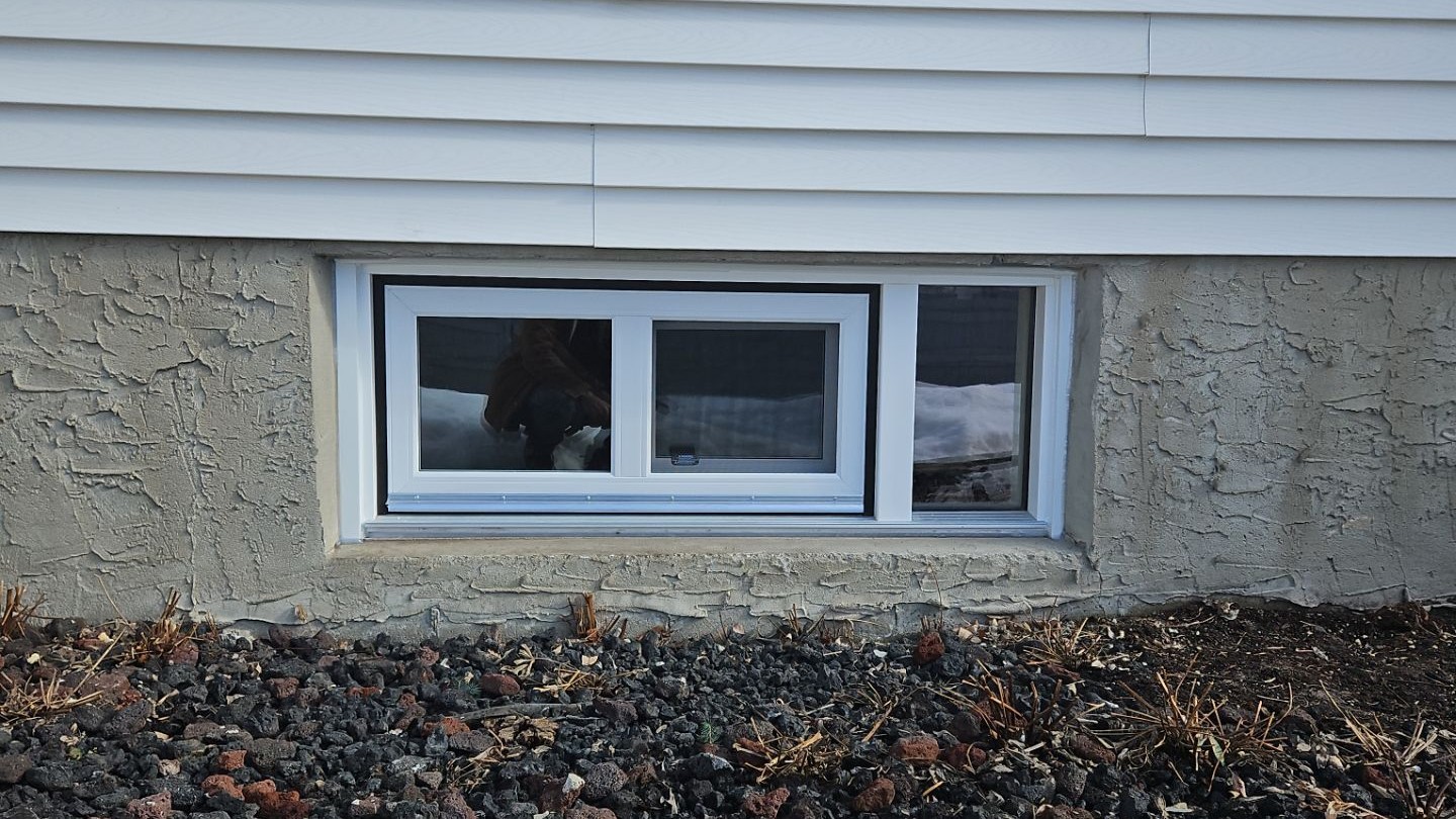 Small basement replacement window with white trim installed in a textured foundation wall beneath light-colored exterior siding.