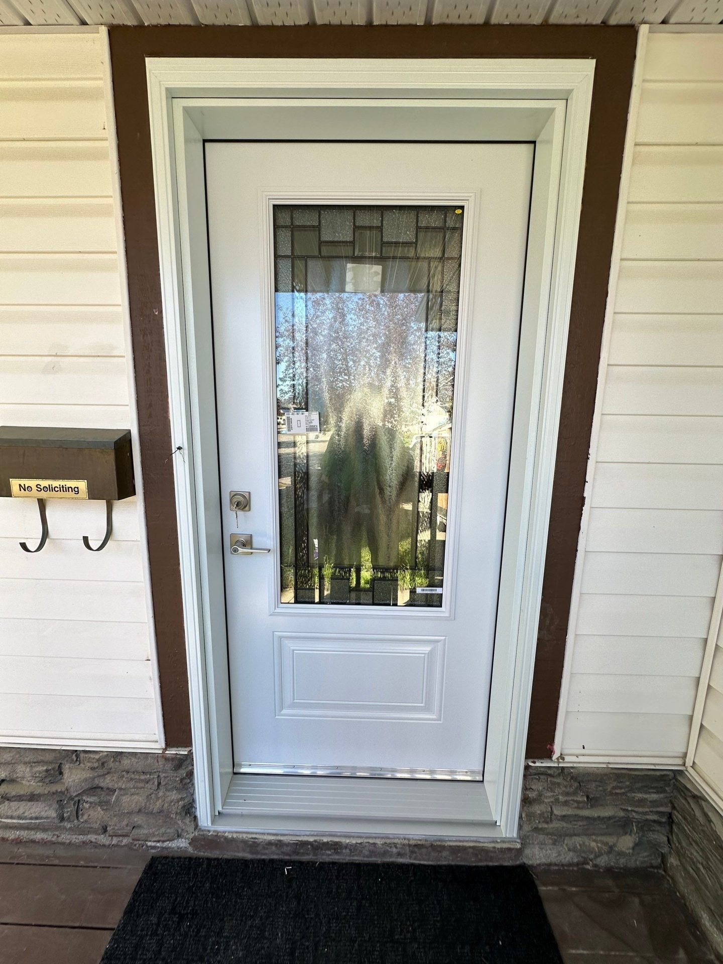 White entry door with decorative glass panel, white trim, cream siding, brown accents, stone base, no-soliciting sign, and black doormat.